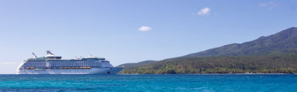 A large cruise ship positioned near a lush tropical island, set against a vibrant blue sky and ocean.